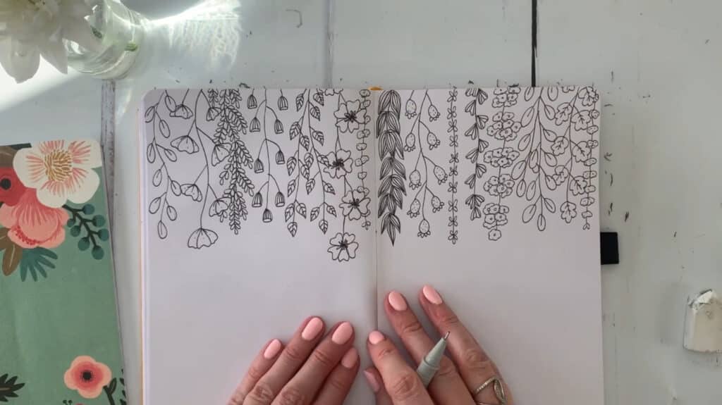 A person’s hands resting on an open sketchbook. It’s filled with doodles of flowers and vines cascading down from the top of the pages. 