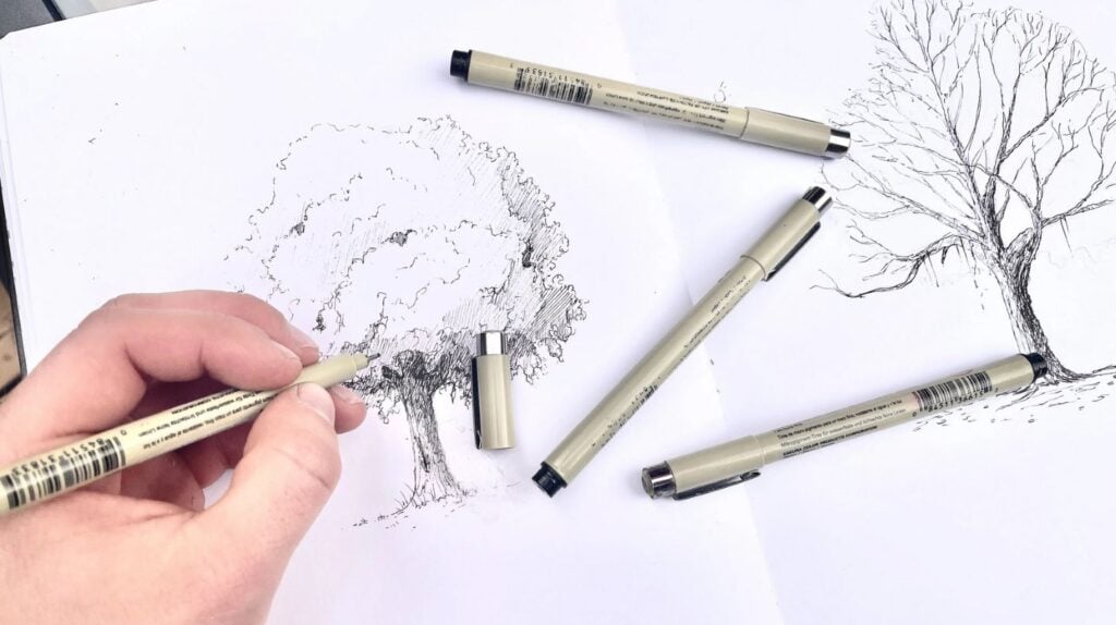 A person’s hand using a pen to draw an illustration of a realistic tree on a white sketchbook page.