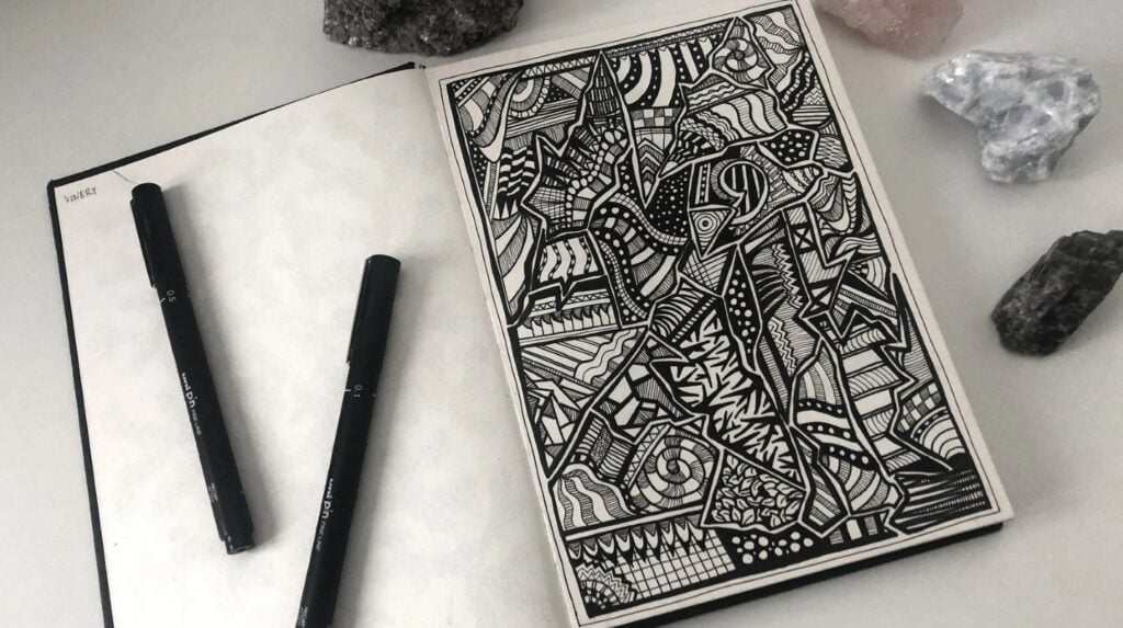 A sketchbook laying open surrounded by crystals. Its page is filled with geometric abstract ink drawings. 