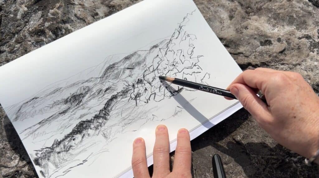 A person’s hand using a pencil to draw a quick sketch of a hilly landscape. Their sketchbook is resting on a rock covered in lichen and moss.