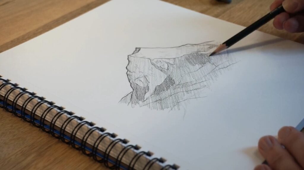 A person using a pencil to create a drawing of a mountain peak in a sketchbook resting on a wooden tabletop.