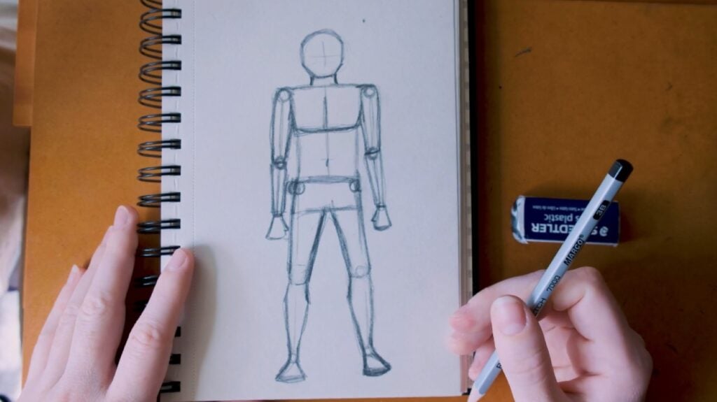 A person’s hand using a pencil to draw an accurately proportioned stick figure on white sketching paper.