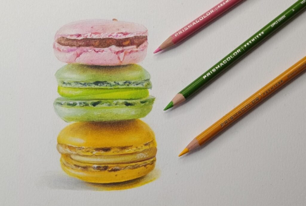 A colored pencil illustration of three macarons stacked on top of each other. One is yellow, one is green and one is pink.