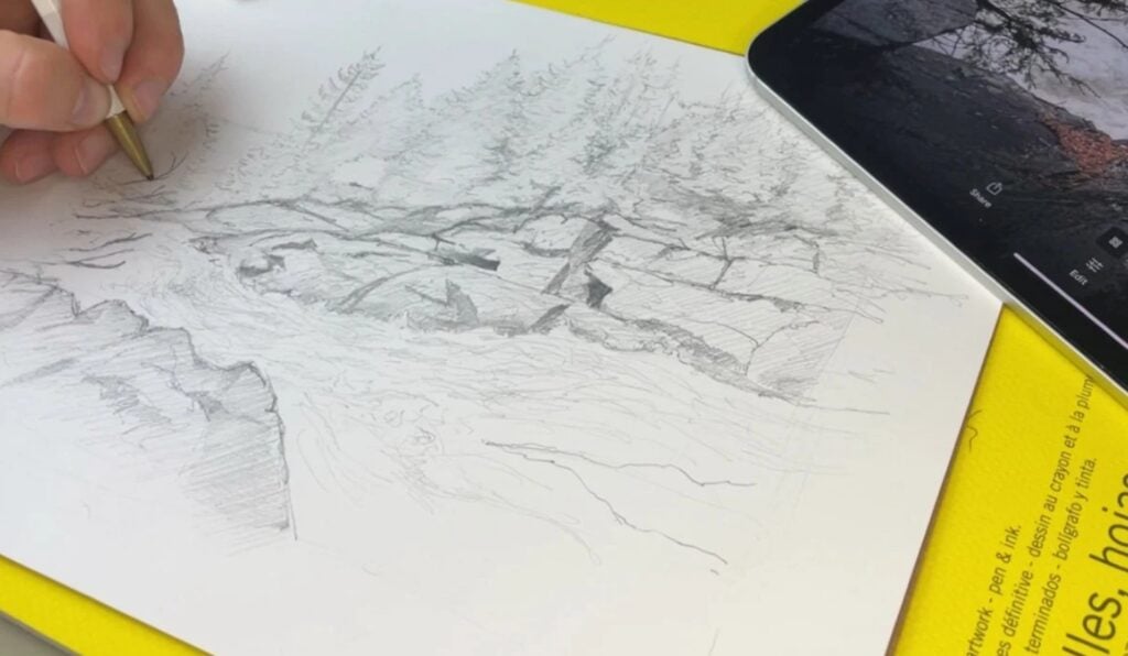 A person’s hand using a pencil to draw a landscape scene of a river rushing over rocks in front of a dense forest.