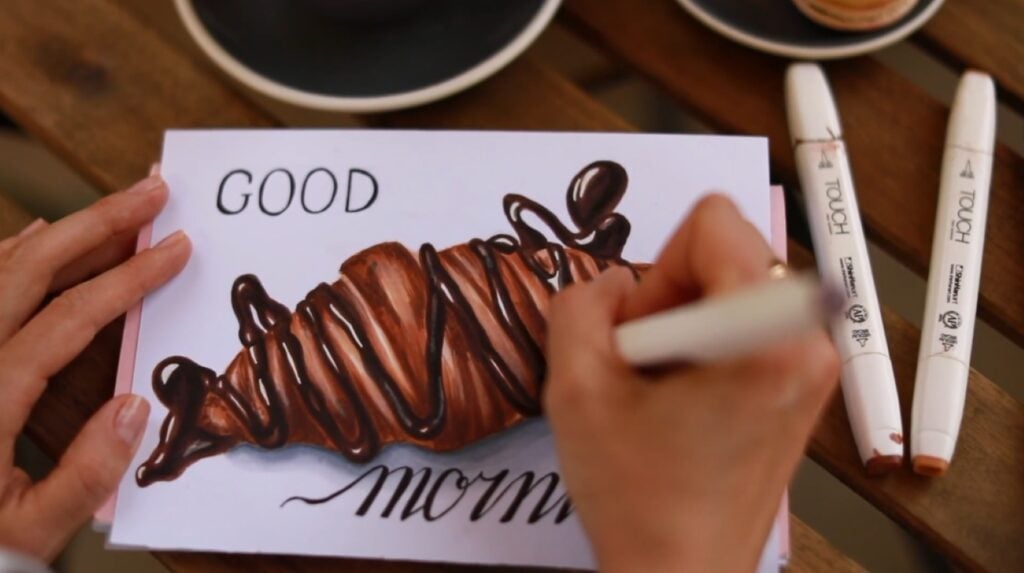 A person using markers to draw a realistic image of a croissant drizzled in chocolate on white paper. 