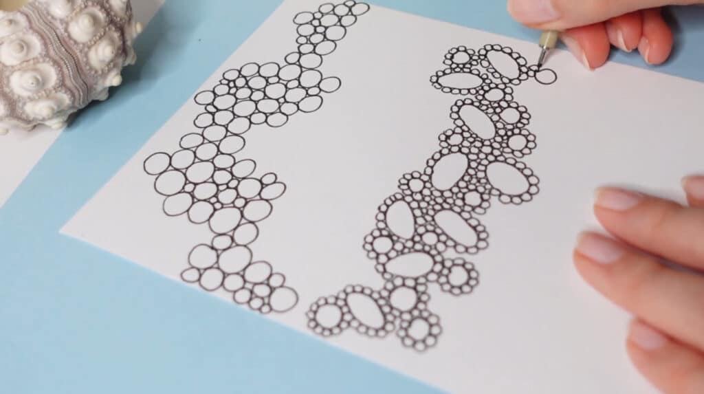 A person’s hand using a pen to draw tiny circles surrounding big circles on a white piece of paper. 