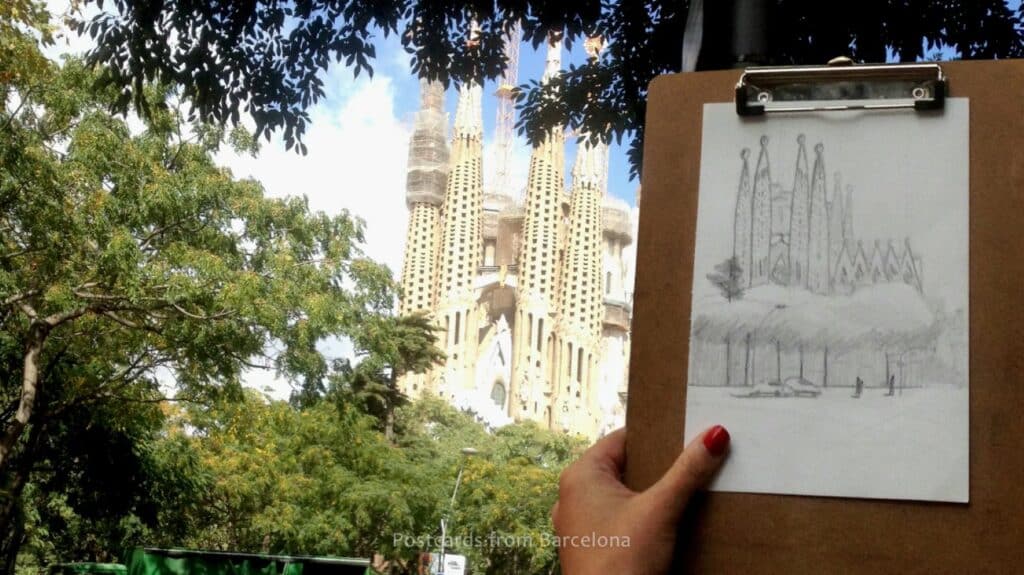 
A person holding up a pencil drawing of a cathedral in front of the cathedral in real life. The drawing is on white paper clipped to a brown clipboard.