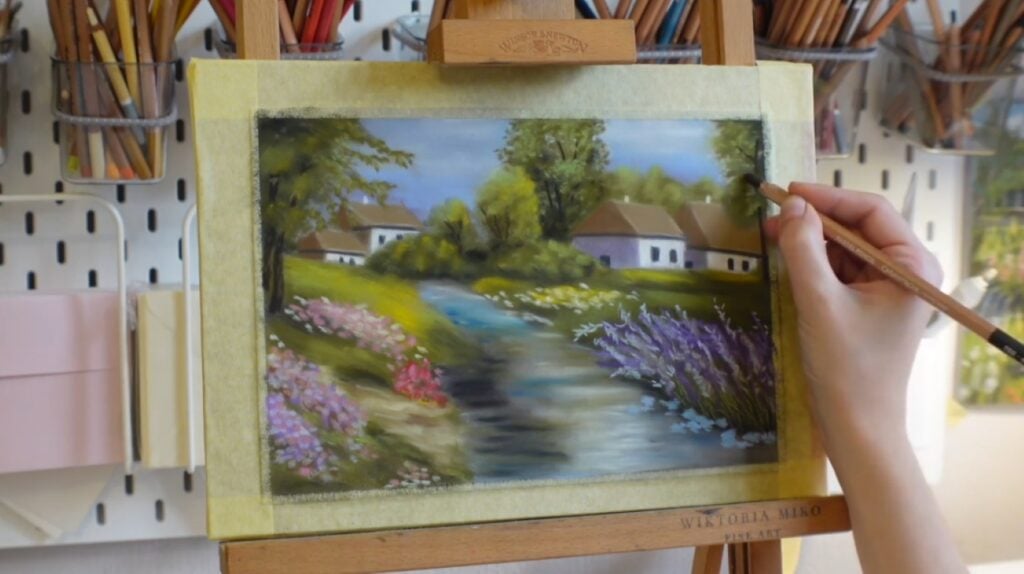 A person’s hand using pastel pencils to create an illustration of idyllic cottages behind a small stream with blooming flowers on either side.