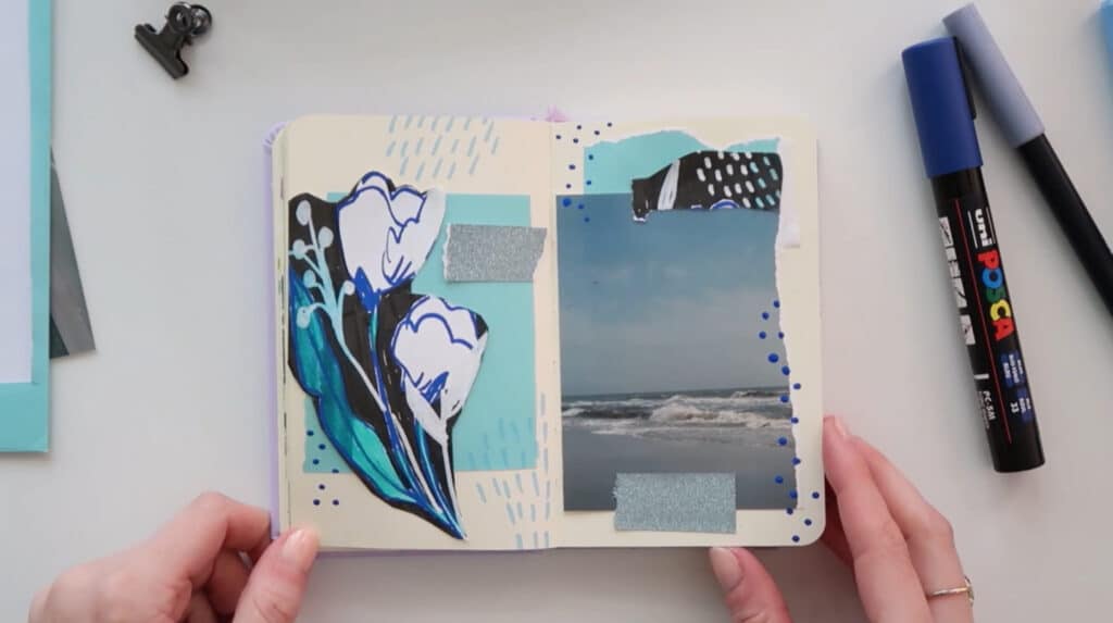 A woman’s hands holding a small notebook open on a white tabletop. The two visible notebook pages have been decorated with a blue-themed collage that includes a photo of the sea under a blue sky, a cut-out sketch of blue flowers, blue scraps of paper and blue doodles.