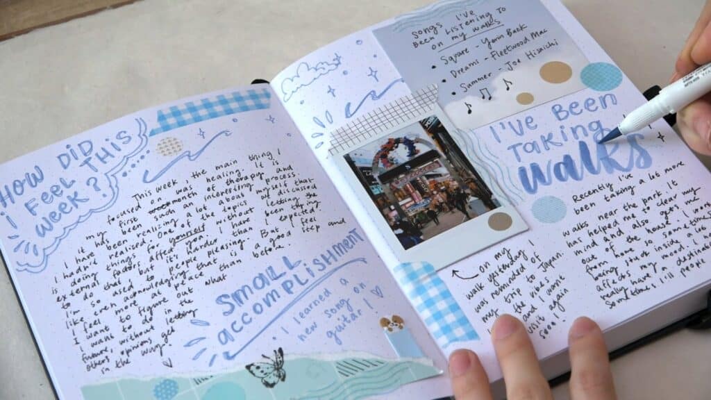 A journal lying open on a cream-colored tabletop. Both pages have been filled with doodles and handwritten text in various shades of blue, as well as a Polaroid photo, scraps of colorful paper, washi tape, stamps and stickers. A woman’s hands can be seen using a calligraphy pen to add blue-colored text.