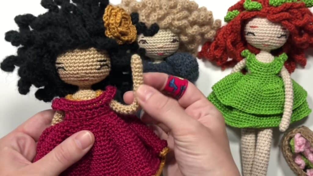 Three amigurumi dolls, one held by the artist with black hair and a wine-colored dress, and two in the background, one with blond hair and a grayish dress, the other with red hair and a green dress