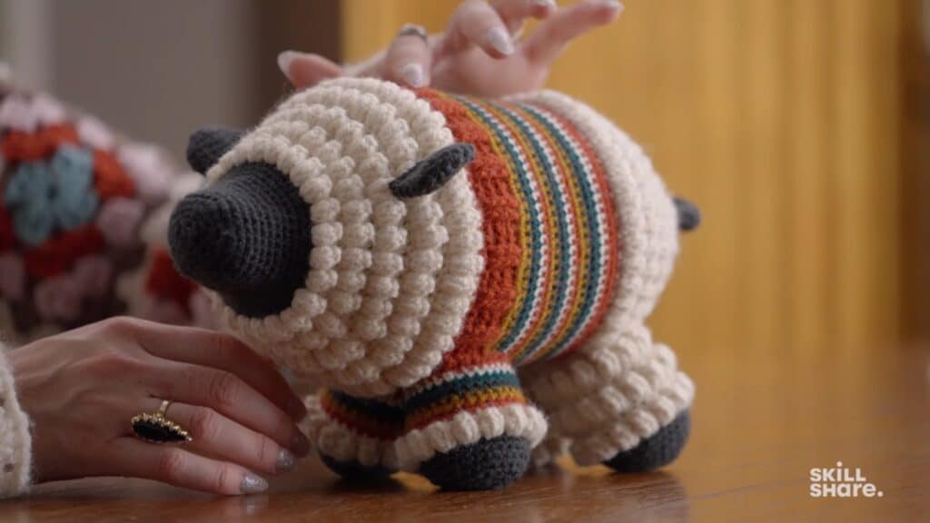 Black and white crocheted sheep wearing a colorful striped sweater and sitting on a table top near the artist’s hands