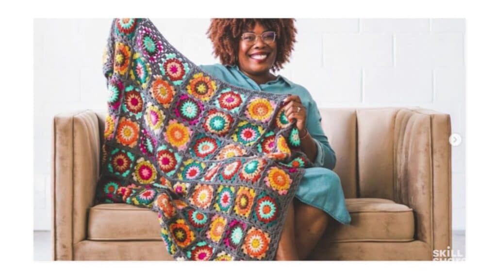 Smiling woman sitting on a tan couch wearing a blue dress and holding up an extremely colorful crocheted blanket