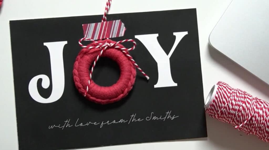 A Christmas card that says ‘joy’ in large white letters laying on a white table. The ‘O’ in ‘joy’ has been replaced with a small red wreath ornament. 