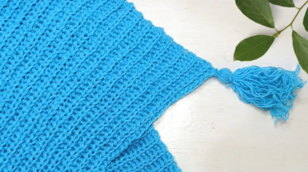 A bright blue crocheted blanket folded on a white tabletop. One of the tassels attached to its corner is visible. 