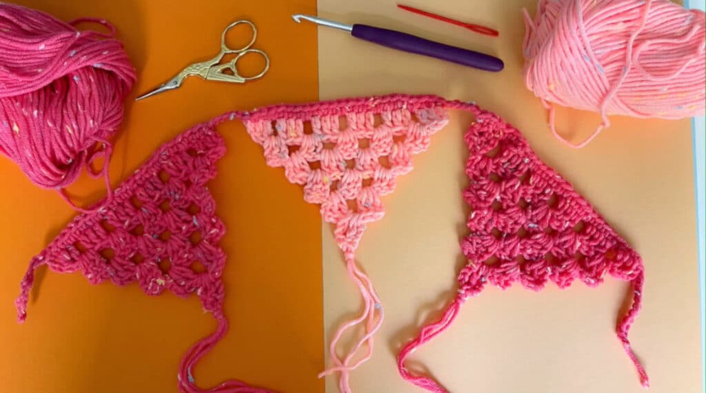 A pink triangle burning garland laying on a tabletop. The garland is crocheted from yarn, and features decorative tassels at the point of each triangle. 