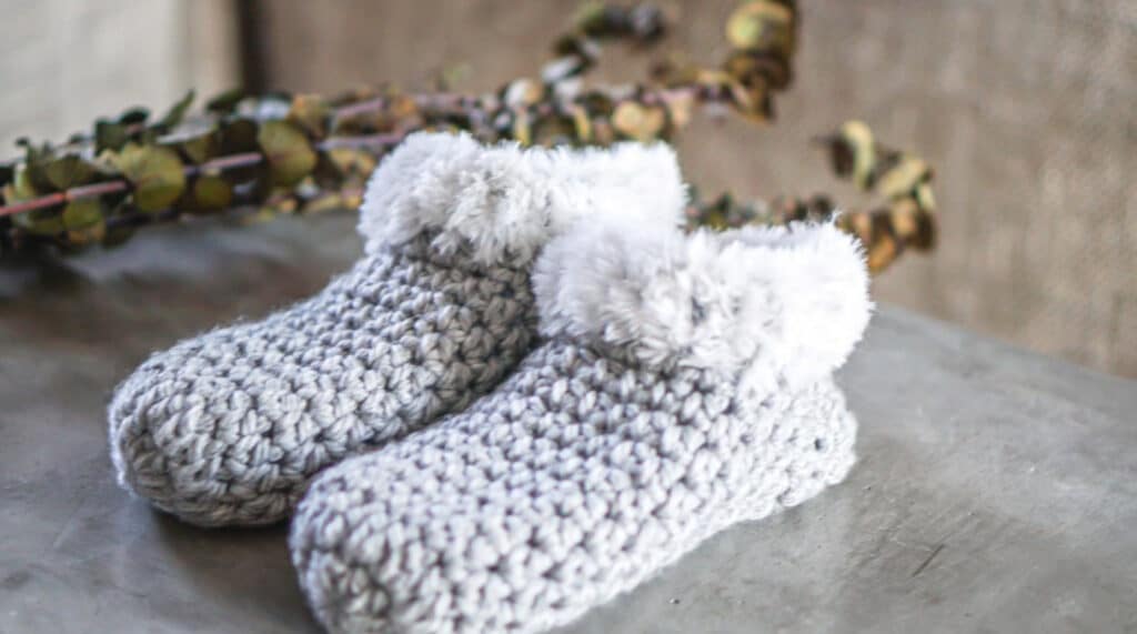 Gray slippers crocheted from chunky yarn and decorated with furry trim around the ankles. They are sitting on a gray tabletop.