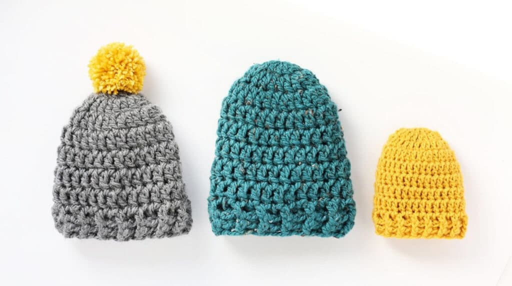Three hats laying out on a white tabletop. One is gray with a yellow pom pom, one is teal with no pom pom and one is small and yellow with no pom pom. 
