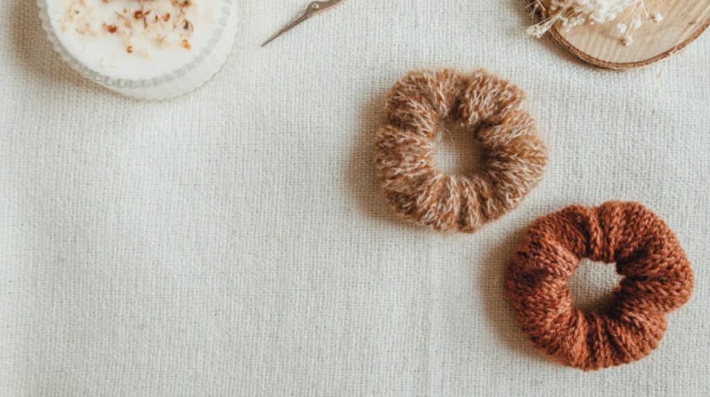 Two knit scrunchies placed on a white cloth surface. One is a dark burnt orange color, and the other is light orange and beige. A white candle in a glass holder is visible in the top left corner, and a wooden coaster is visible in the top right corner.