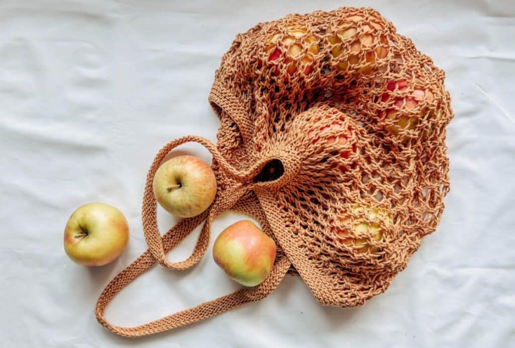 A light orange knit lace tote bag lying on a white cloth surface. The bag has been filled with apples, and three have rolled out onto the cloth-covered tabletop.