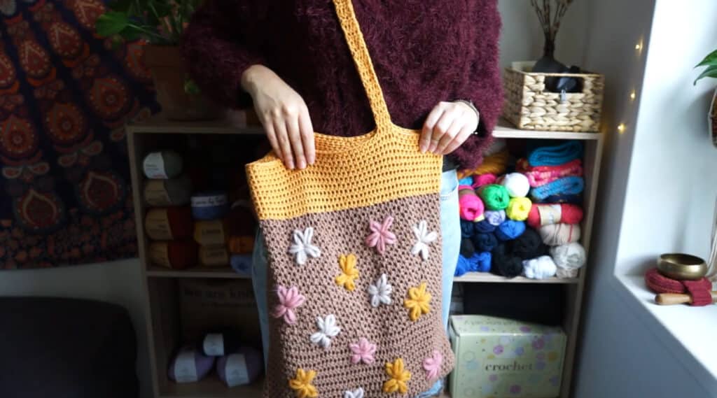 A woman wearing a brown tote bag with a light orange top section and straps. The bag is decorated with embroidered white, pink and orange flowers. 