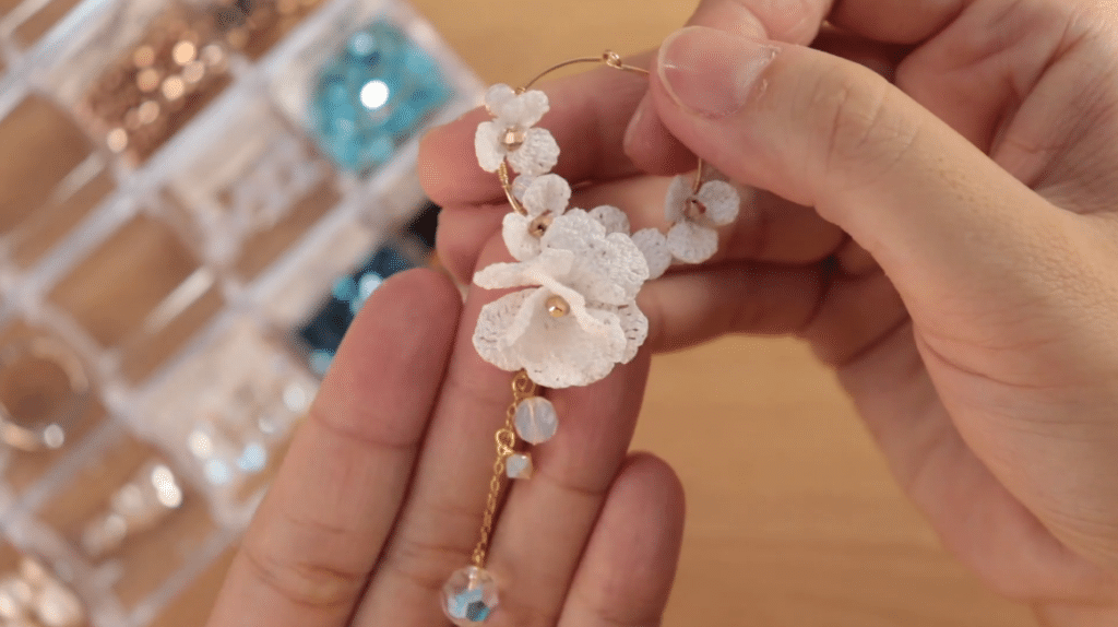 A person’s hands holding a gold hoop earring that’s been decorated with white crocheted flowers, a gold chain and sparkling crystal charms. 