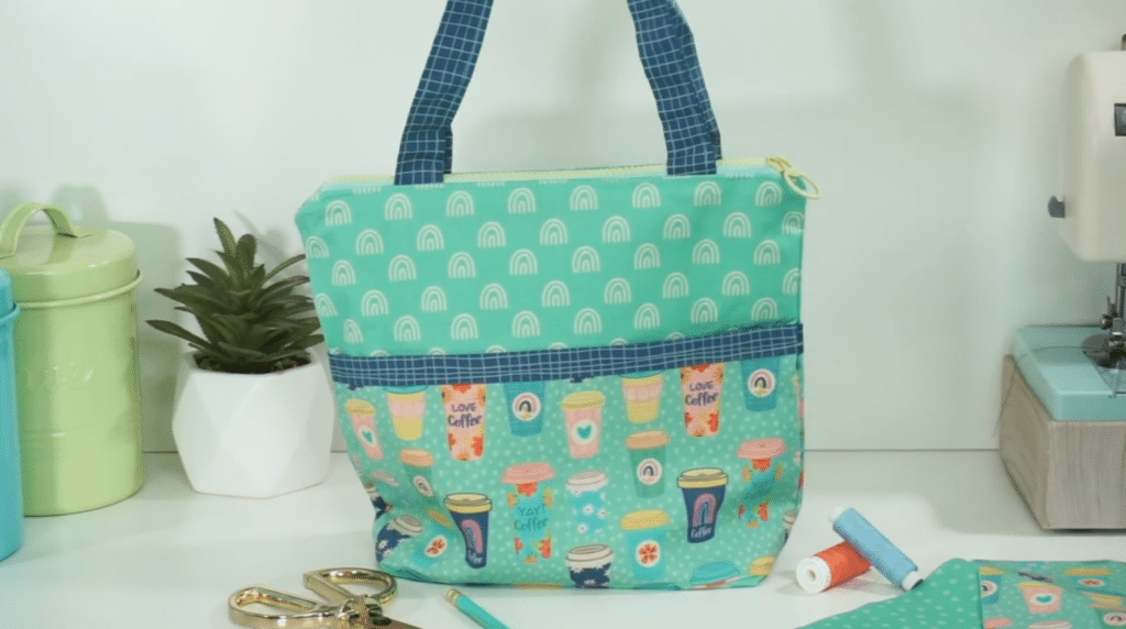 A tote bag sits on a white table alongside a potted succulent, a pair of scissors, thread, a pencil, fabric and a sewing machine. The tote bag has navy blue and white striped handles. The main part of the bag is made up of two different light green fabrics. One fabric is covered in white rainbows and the other in colorful, to-go coffee cups.
