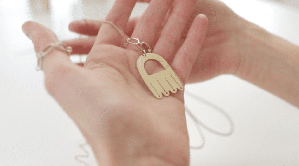 Two hands hold a gold necklace. The necklace has a thin gold chain and a big, upside-down U-shaped charm. 