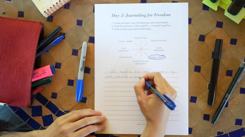 A woman’s hands writing on a piece of paper lying on a tiled tabletop. The paper is a worksheet titled ‘Day 3: Journaling for Freedom.’ It instructs students to circle an area in their life where they want more freedom, then write a journal entry that explains how they’ll give themselves that freedom.