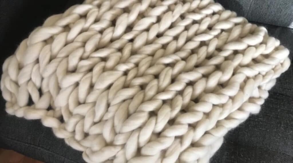 A white chunky knit blanket folded in a square and placed upon a gray upholstered couch. The blanket is made of fluffy strands of unspun wool.