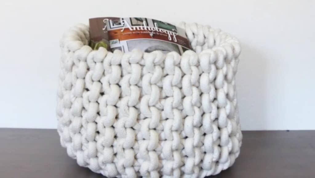 A white knit storage basket sitting on dark flooring in front of a white wall. The basket is made from thick cotton yarn. The basket is holding copies of a magazine titled ‘Anthology.’