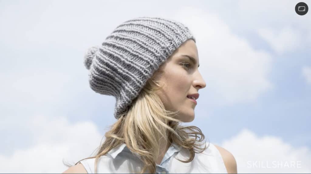 A woman standing underneath a blue sky with white clouds, wearing a gray ribbed knit beanie that’s decorated with a pom-pom on the top.