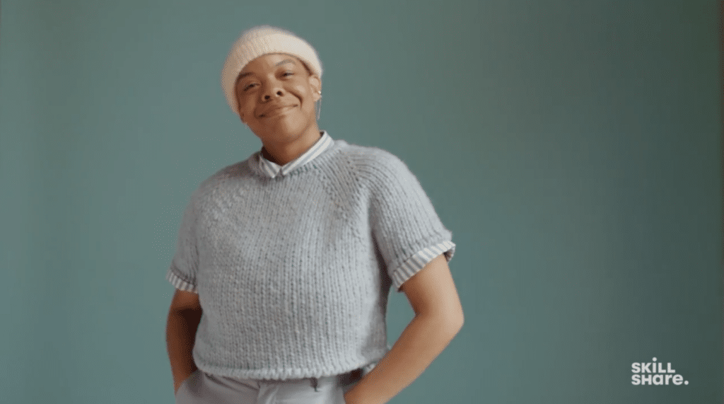 A person wears a cream, knitted cap and a light gray short-sleeved knit sweater. The person is smiling and standing against a dark gray background. 