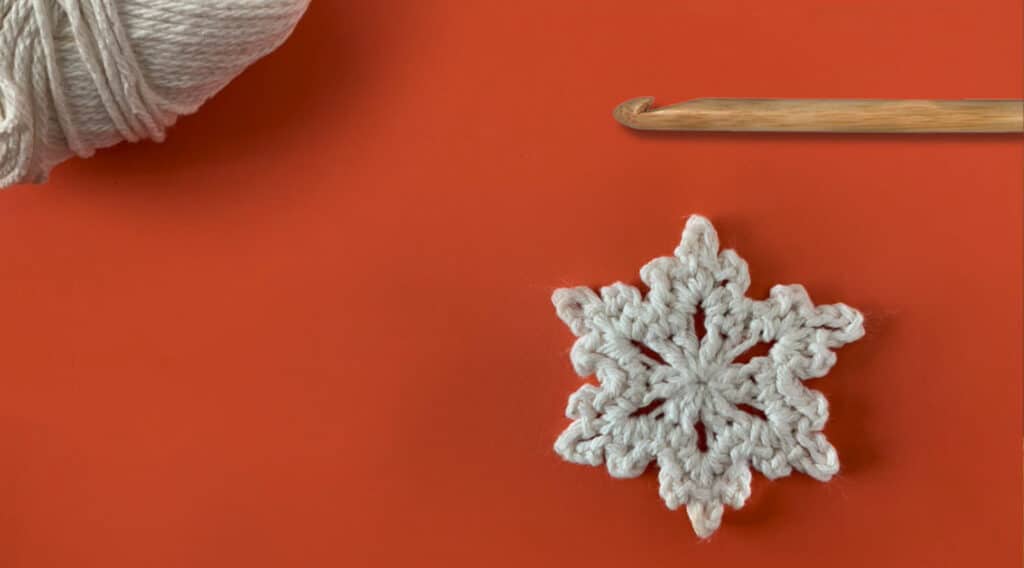A white crocheted snowflake and a wooden crochet hook laying next to each other on a red tabletop.