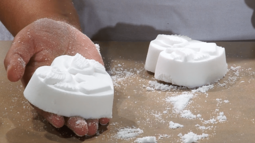 A hand holds a heart-shaped, white bath bomb over a wooden table. The bath bomb has winged creatures and a heart imprinted on it. In the background, sitting on the wooden table, there is another identical bath bomb. 