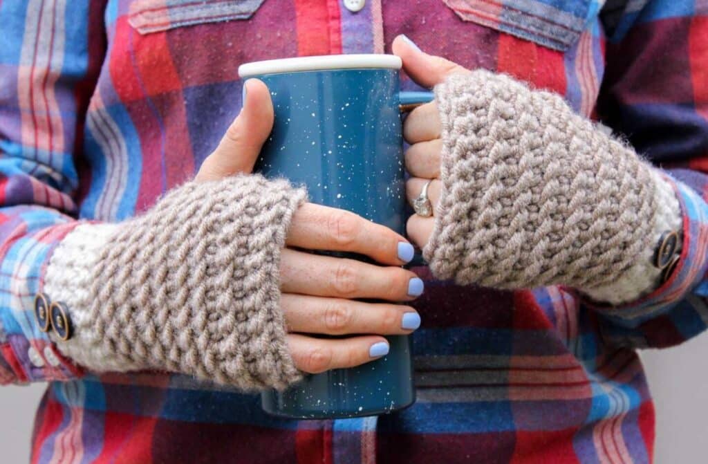 A person’s hands wearing taupe crocheted fingerless gloves while holding a green speckled travel coffee mug. 