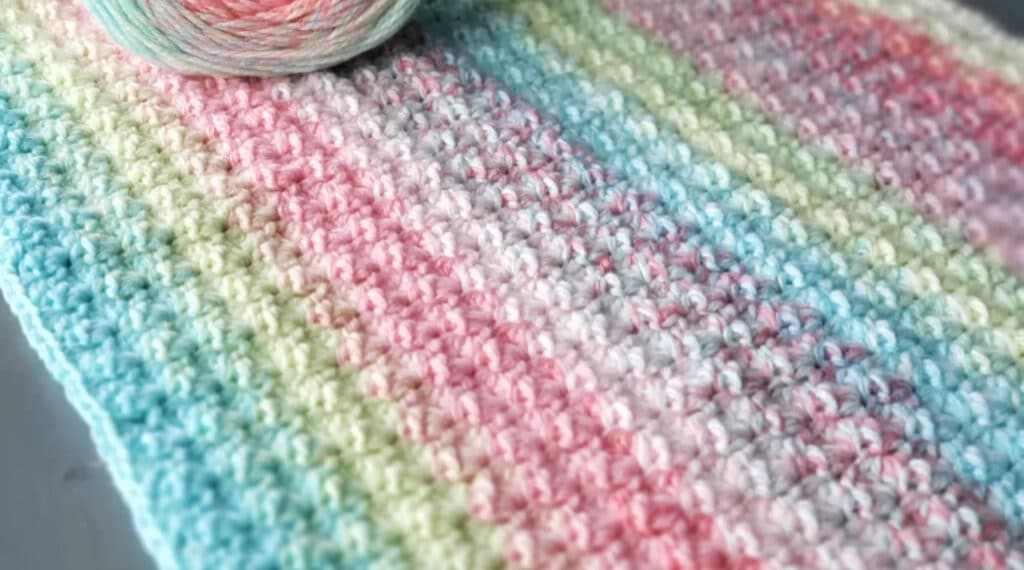 A crocheted blanket in pastel shades of blue, green, pink and white laid out on a flat surface.