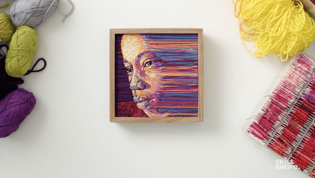 An embroidered work of art sits in a wooden frame on a white background surrounded by different colored skeins of yarn and thread. The work of art features a woman's face looking into the distance. Her face is made up of different colored yarn and threads and then transitions into straight lines of yarn and thread. 