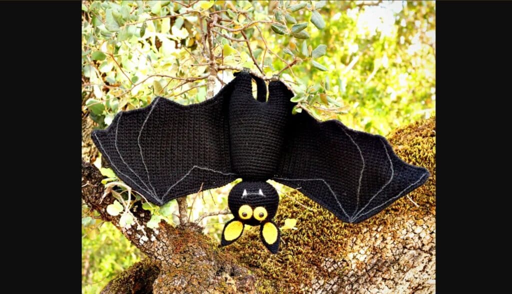 Crocheted black bat with yellow eyes and inner ears, hanging upside down from a branch in a real tree