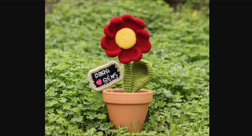 Crocheted flower with red, heart-shaped petals, yellow center, green stem and leaf in a terracotta pot, planted next to a little crocheted sign reading “Para Bens” with a pink heart, all surrounded by clovers and grasses