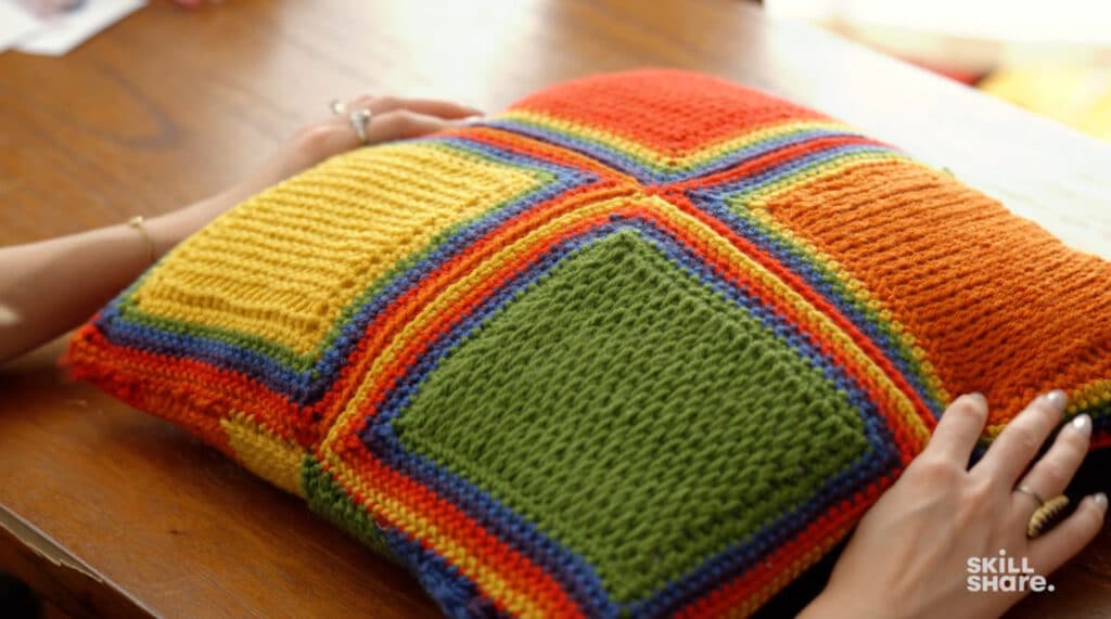 A person’s hands holding a pillow on a wooden tabletop. The pillow is crocheted and features vibrant squares of green, orange, yellow, red and blue.