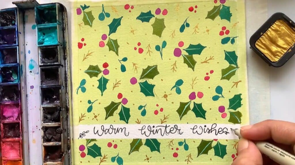 A person’s hand using a pen to write the words ‘warm winter wishes’ in cursive script on a greeting card. The card is light green and features dark green holly leaves and red berries painted in a pattern.
