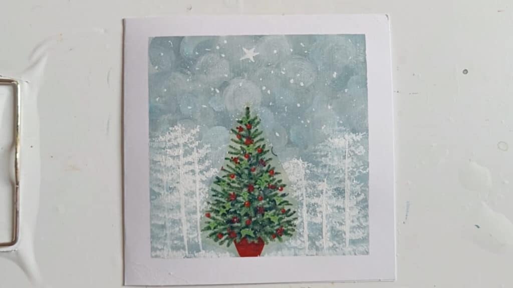 A square white greeting card laying on a flat white surface. The card is painted with a depiction of a green Christmas tree decorated with red ornaments that’s standing in a blue and white toned forest under a starry sky.