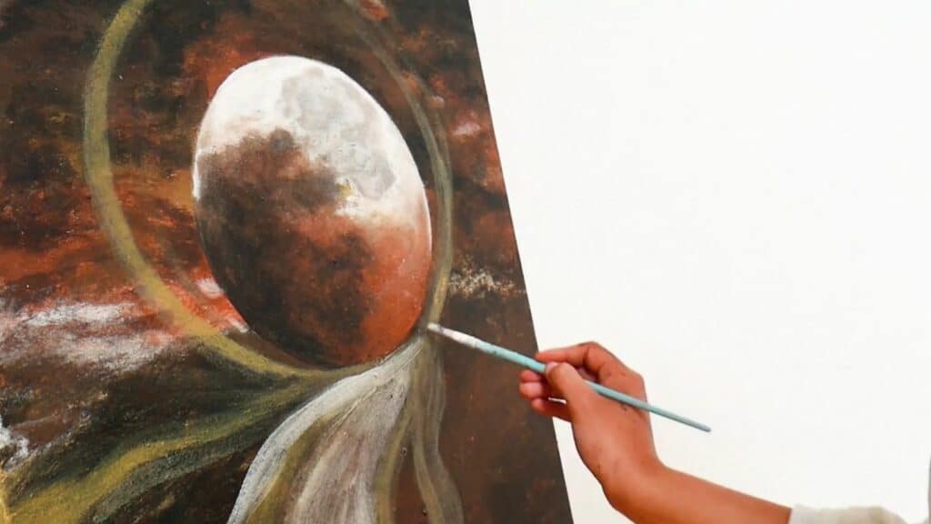 Artist’s hand applying a brush to a canvas, on which there’s an abstract painting of a planetary setting in reddish browns, greens, blacks and whites