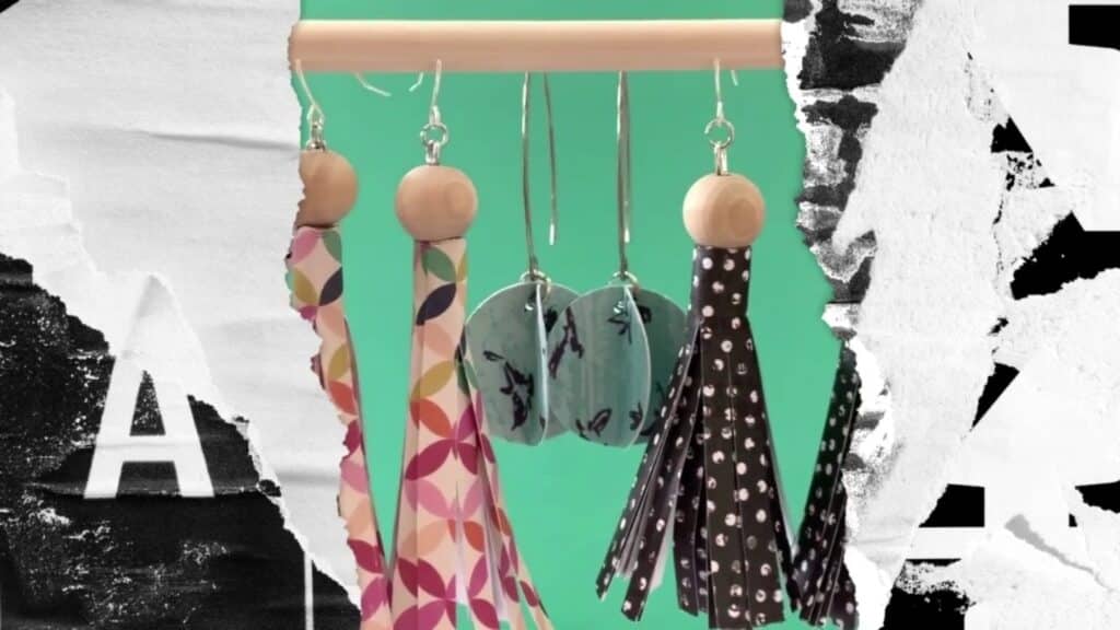 Three pairs of paper earrings, in grays, pinks, blacks, and silvers, hanging from a wooden dowel; on either side is a torn paper motif in black and white 