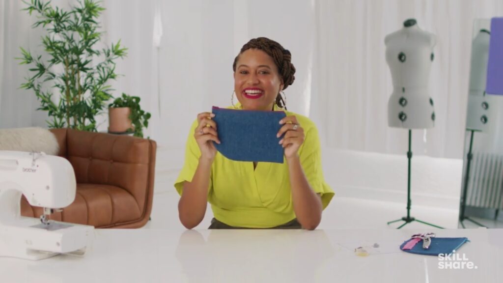 Smiling woman holding up a medium-sized blue zippered bag, sitting at a white work table against the background of a white wall and curtains, plants, brown leather cough and dress form 