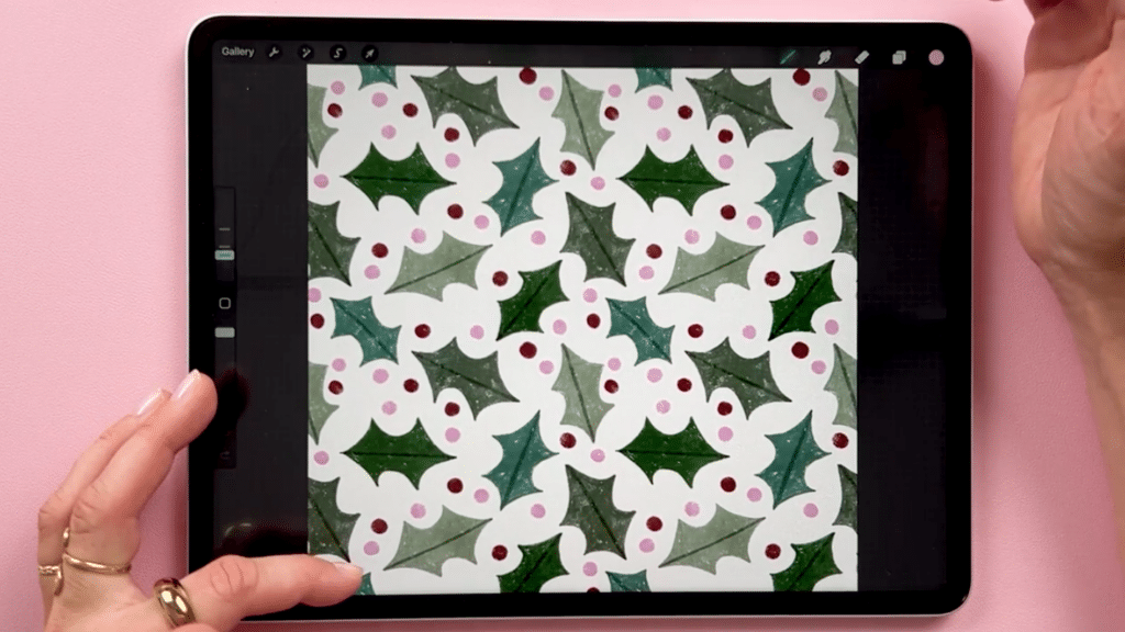 An iPad sits on a pink background. Two hands sit to each side with one touching the screen in the bottom left corner. The iPad is open to Procreate where there is an illustrated pattern of a bunch of holly leaves and red and pink polka dots.