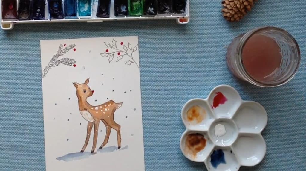 A white greeting card laying on a blue cloth tabletop. The card is hand-painted with a brown and white spotted fawn that’s standing in a snowy forest. A small palette of watercolors is visible to the right of the card.
