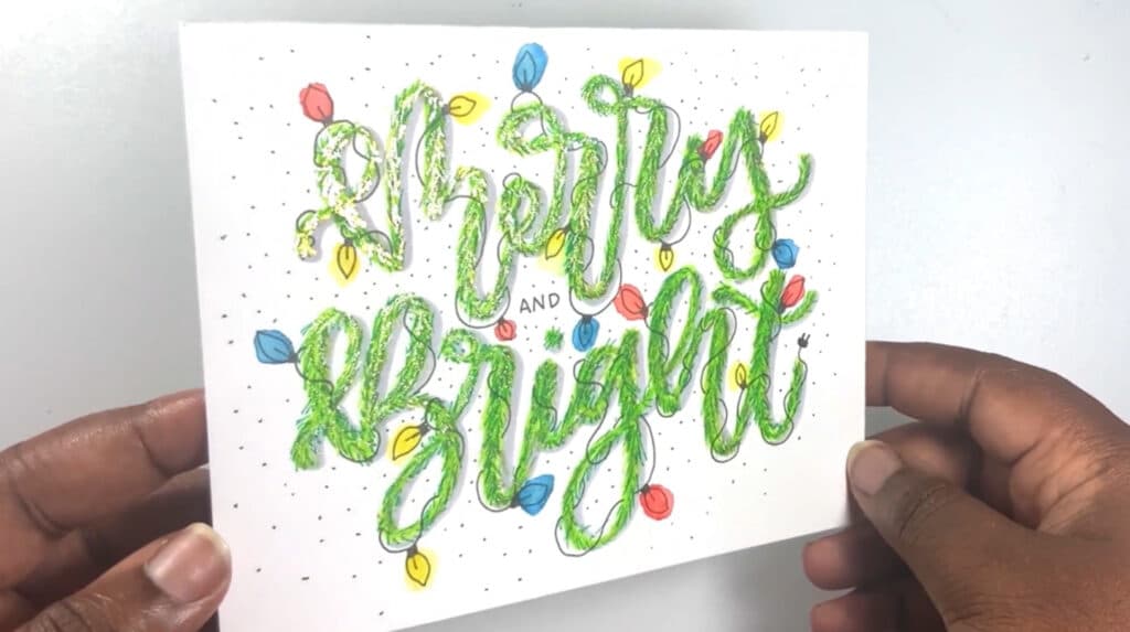 A person’s hands holding a white greeting card that reads ‘merry and bright’ in hand-lettered text that looks like it’s made from a pine garland. The text is also decorated with colorful illustrated Christmas lights, and the letters are adorned with gold accents.