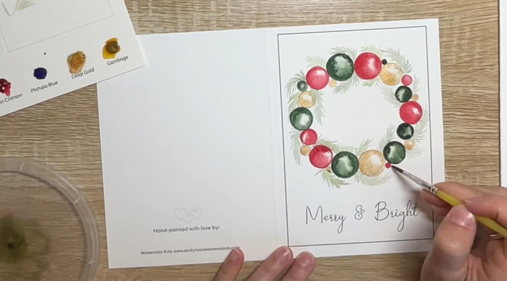 A person’s hands using a paintbrush to paint a greeting card. The greeting card features a wreath made of ornaments and pine boughs, with the text ‘merry and bright’ written underneath it.